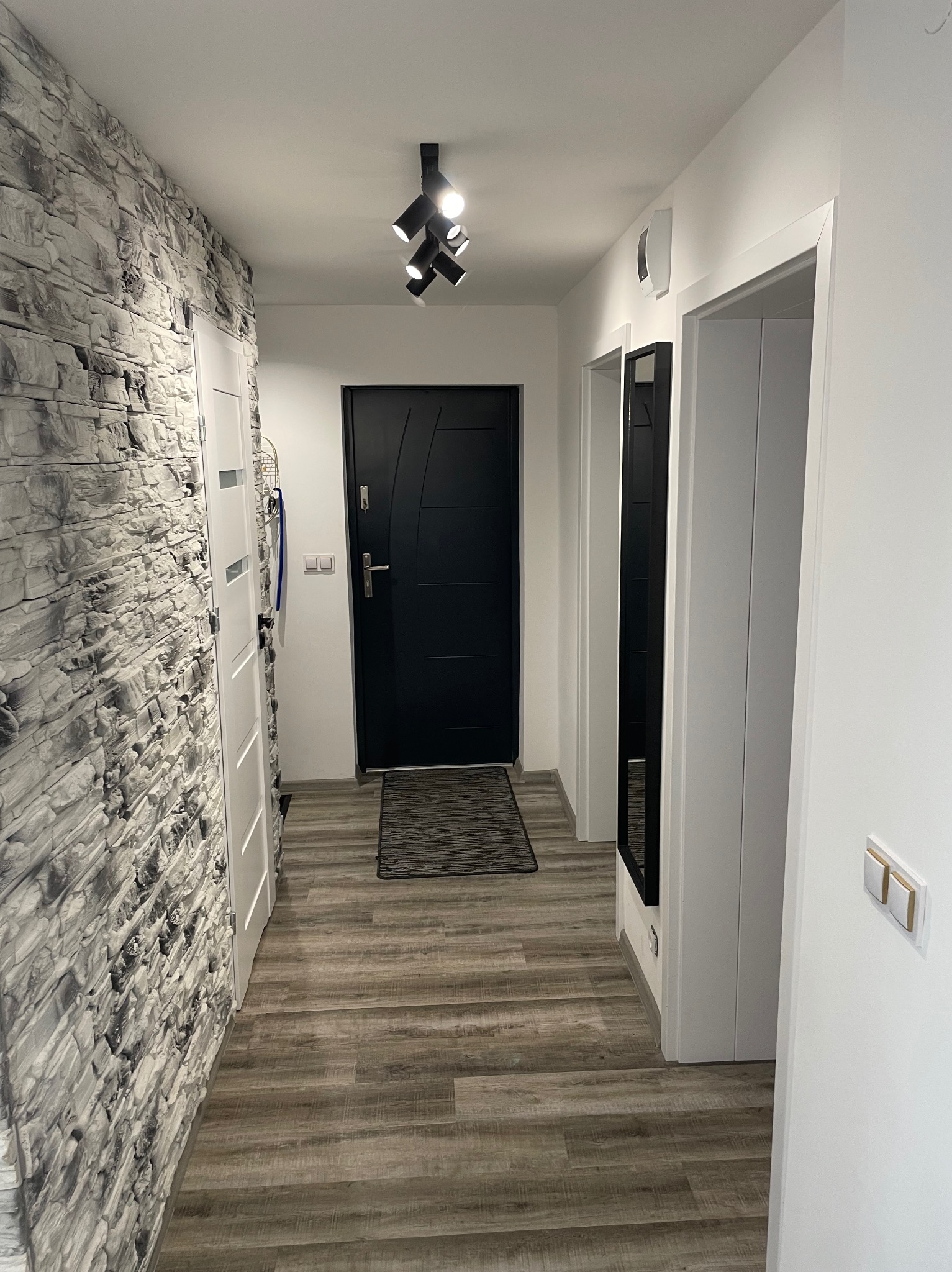 Nowoczesny korytarz z kamienną ścianą, panelami podłogowymi w odcieniach szarości, czarnymi drzwiami wejściowymi i punktowym oświetleniem sufitowym.