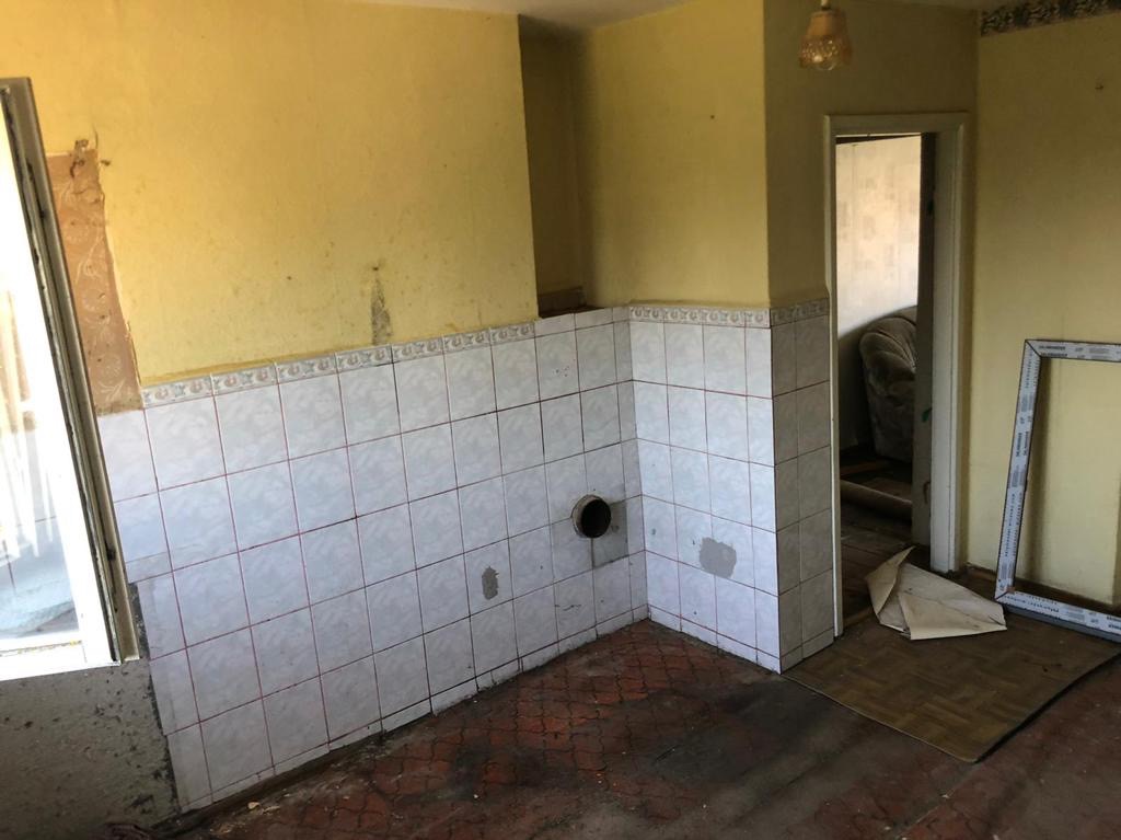 Żółta ściana z częściowo położonymi białymi płytkami z bordiurą, widoczne ślady po usuniętych elementach i otwór w ścianie, podłoga z resztkami starej wykładziny.