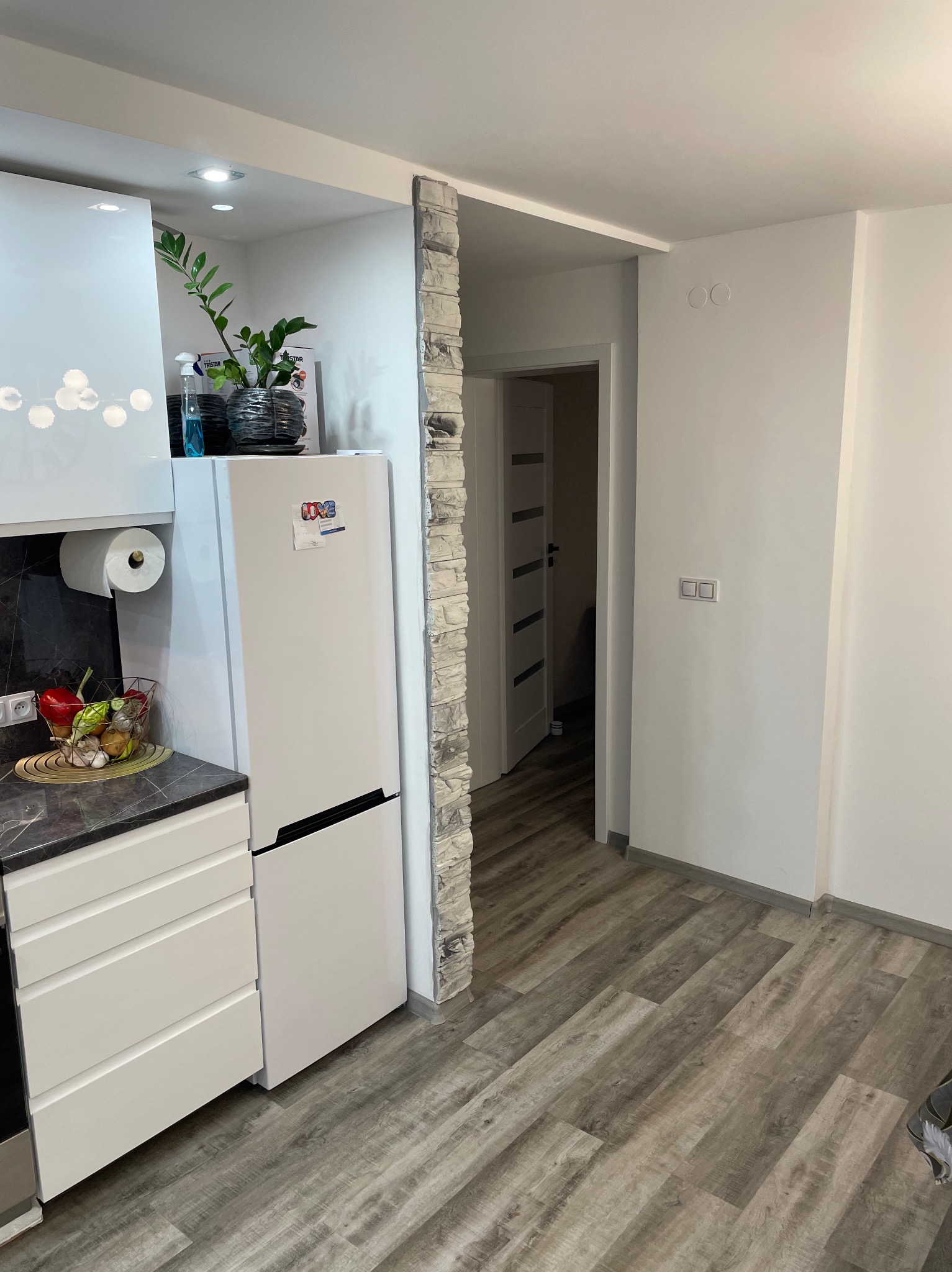 Nowoczesna kuchnia z białymi meblami, marmurowym blatem, drewnopodobną podłogą i dekoracyjną ścianą z imitacji kamienia przy przejściu do innego pomieszczenia.