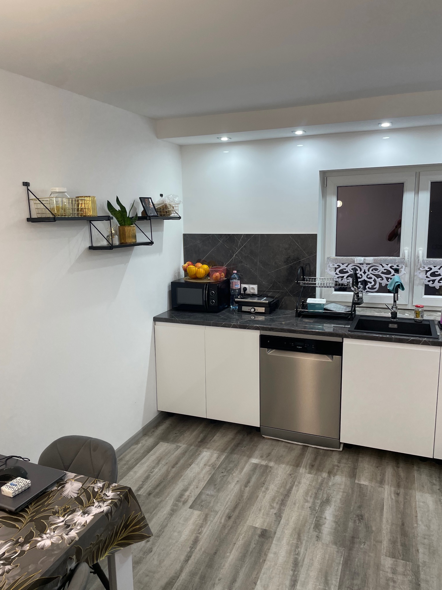 Nowoczesna kuchnia z białymi szafkami, szarą ścianą nad blatem imitującą marmur, szarą podłogą imitującą drewno i czarnymi metalowymi półkami z dekoracjami.