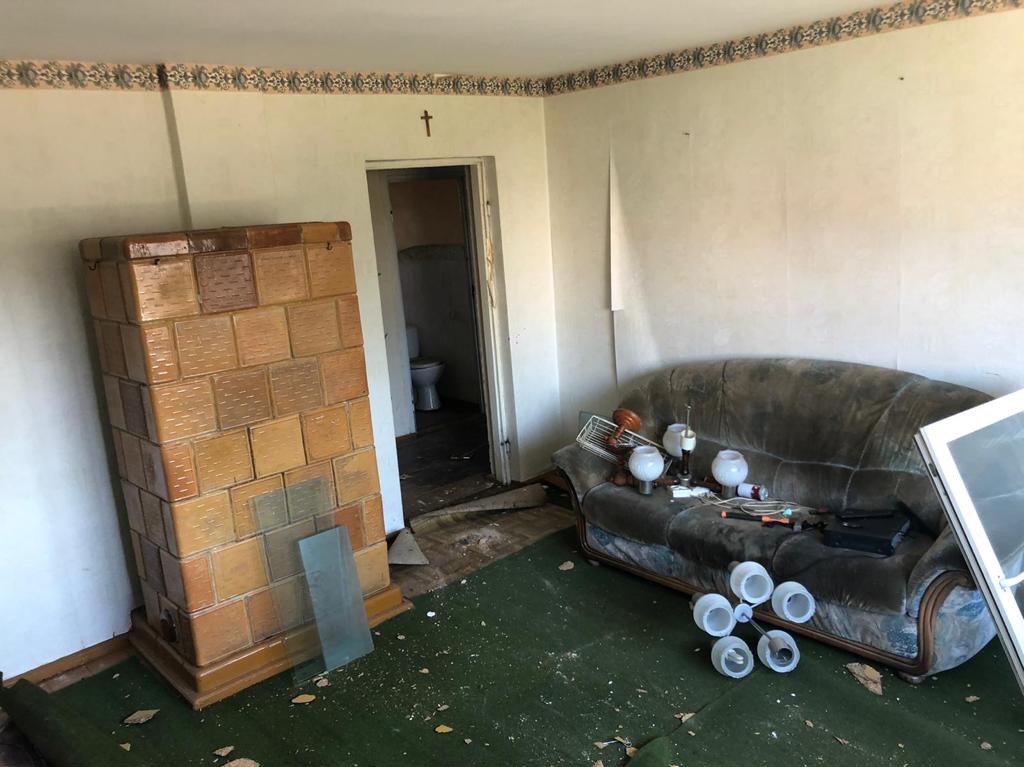 Pokój w trakcie demontażu: stary piec kaflowy, zniszczona sofa z lampami, odpadająca tapeta, widoczny fragment łazienki z toaletą w tle.