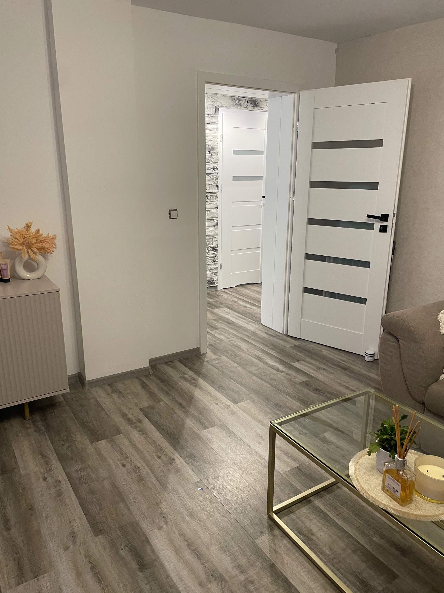 Nowoczesne wnętrze mieszkania z podłogą imitującą drewno, białymi drzwiami z poziomymi przeszkleniami i szklanym stolikiem kawowym z dekoracjami.