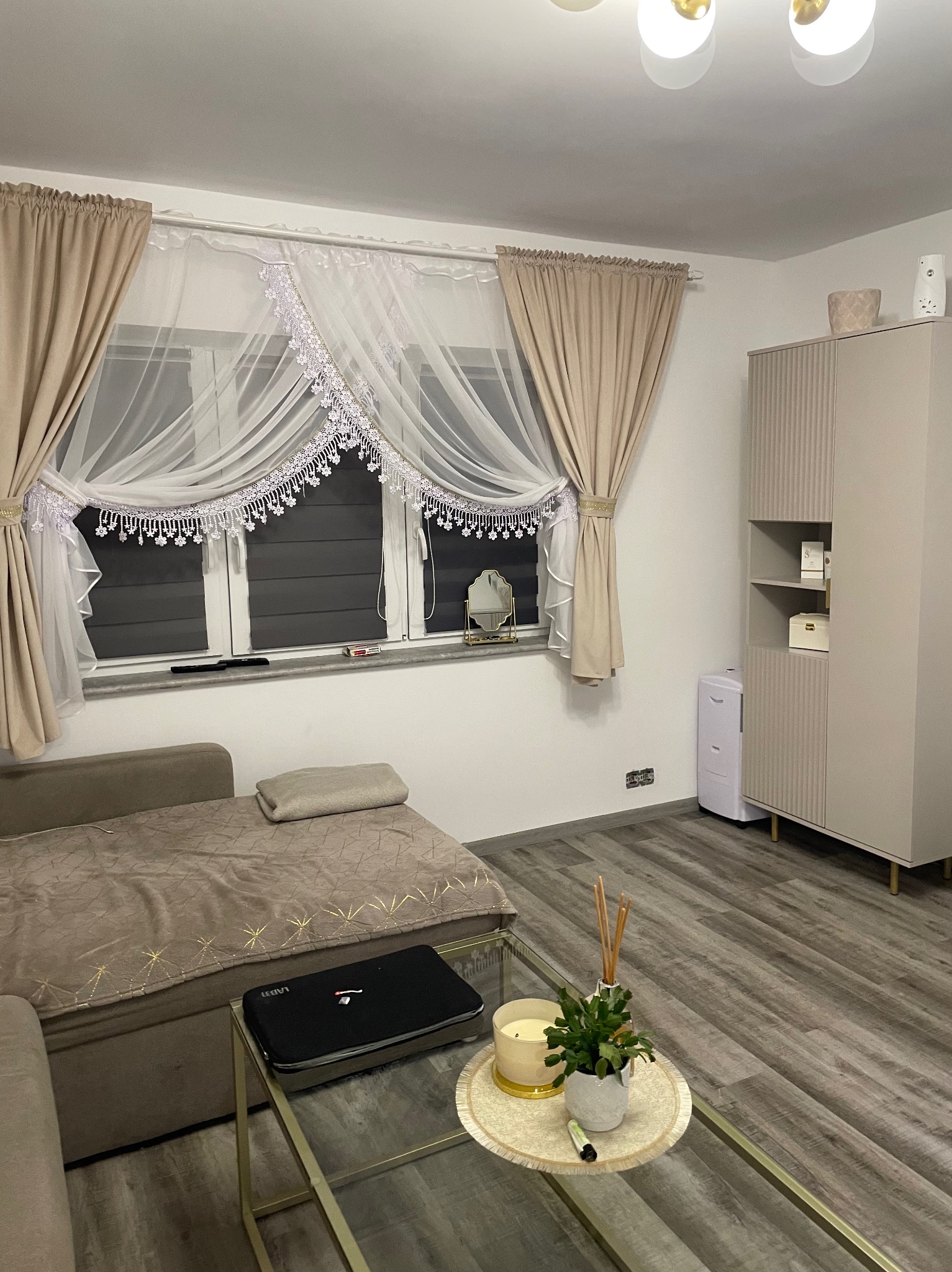 Beżowo-białe zasłony z koronkowym wykończeniem w oknie, szara podłoga, sofa w odcieniach brązu z narzutą w geometryczne wzory, szklany stolik kawowy z dekoracjami, szafa w kolorze ecru z półkami...