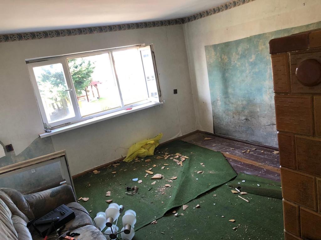 Pokój w trakcie demontażu elementów wykończeniowych: zerwana zielona wykładzina dywanowa, częściowo odsłonięta drewniana podłoga, odsłonięte fragmenty ścian, widoczny stary piec kaflowy, okno...