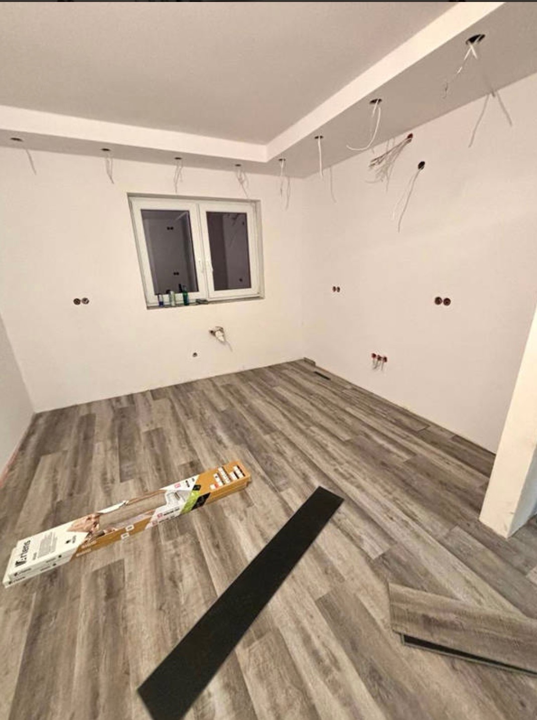 Wnętrze pokoju w trakcie układania paneli podłogowych, widoczne paczki paneli oraz częściowo ułożona podłoga w odcieniach szarości, na ścianach widoczne wyprowadzenia elektryczne.