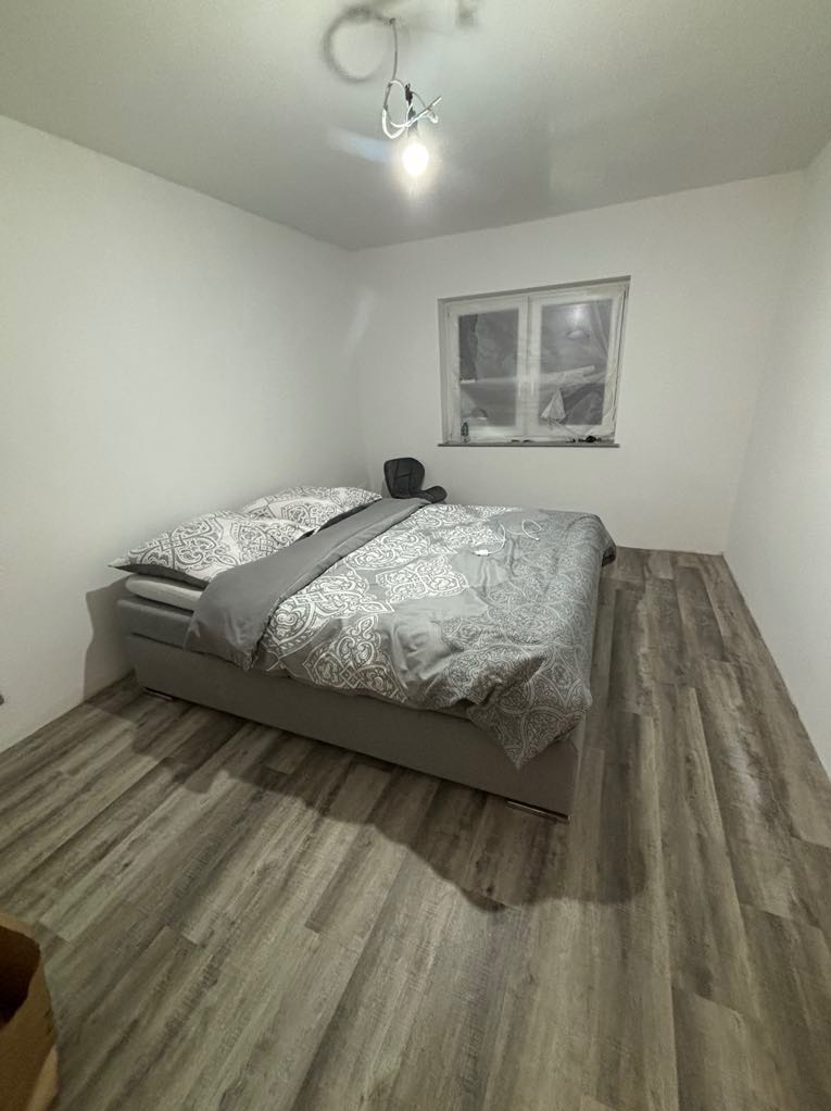 Pokój z nową podłogą z paneli w odcieniach szarości, łóżko z szarą pościelą, okno zakryte folią malarską, surowa żarówka na suficie.