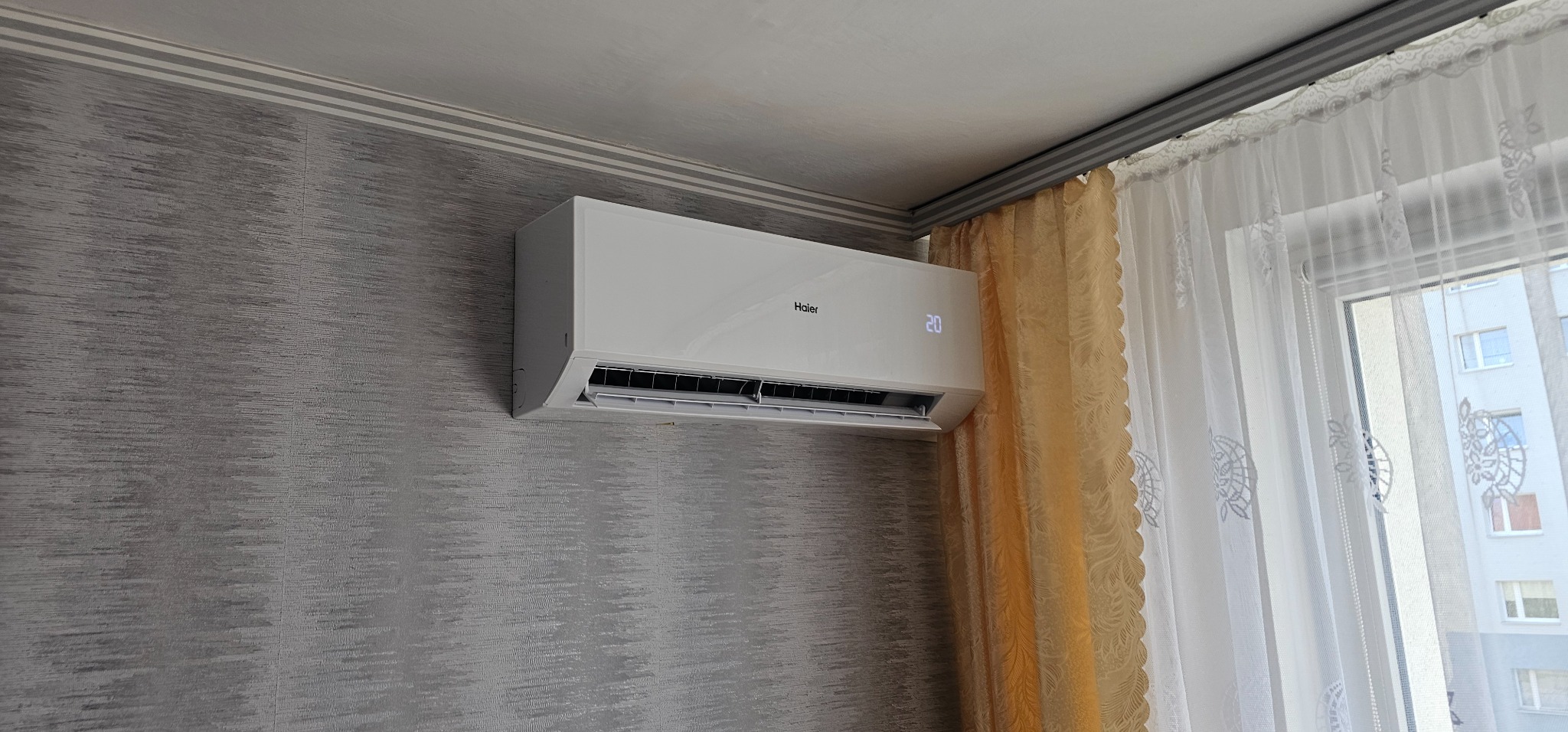 Biały klimatyzator Haier zamontowany na szarej ścianie z tapetą w pokoju z beżowo-białymi zasłonami i oknem.