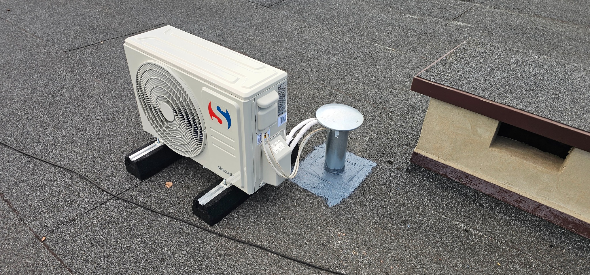 Zewnętrzna jednostka klimatyzacji Sinclair zamontowana na płaskim dachu pokrytym papą, widoczne przewody i elementy montażowe oraz komin wentylacyjny.