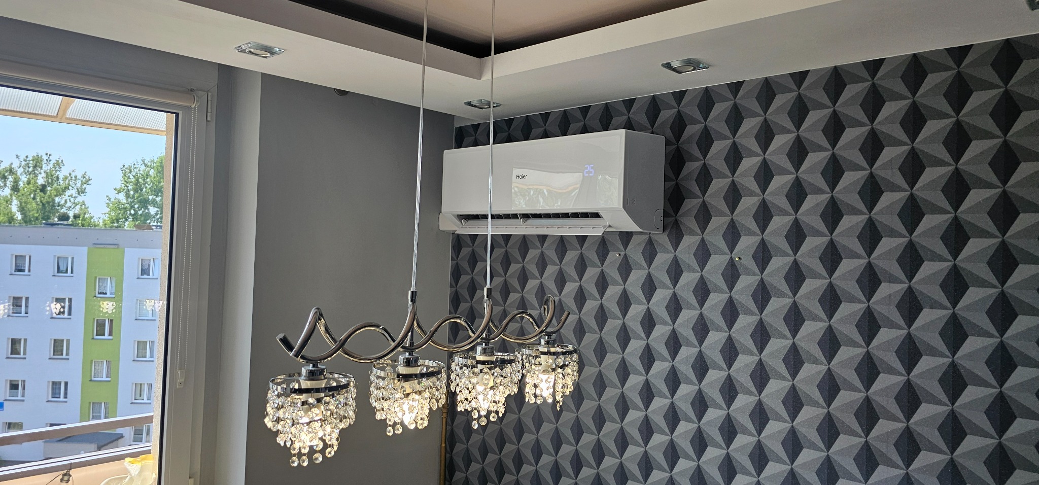 Biały klimatyzator Haier zamontowany na ścianie z trójwymiarową tapetą w pokoju z oknem i ozdobnym żyrandolem.