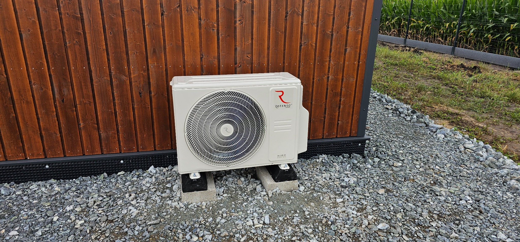 Zewnętrzna jednostka klimatyzacji Rotenso Hiro Series zamontowana na betonowych podstawach przy drewnianej ścianie budynku, otoczona żwirem.