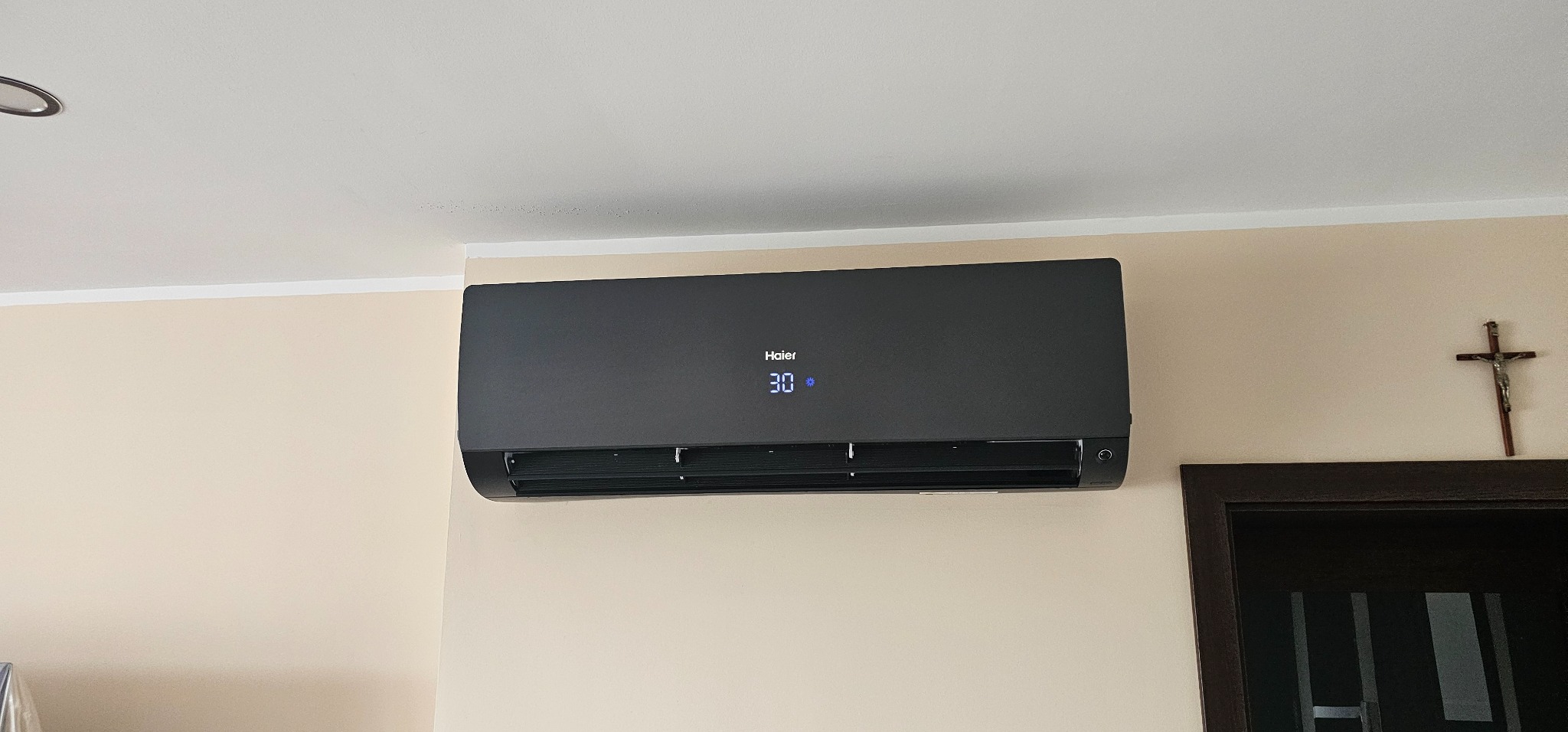 Czarny klimatyzator Haier zamontowany na beżowej ścianie, wyświetlający temperaturę 30 stopni, obok futryny drzwi i krzyża.