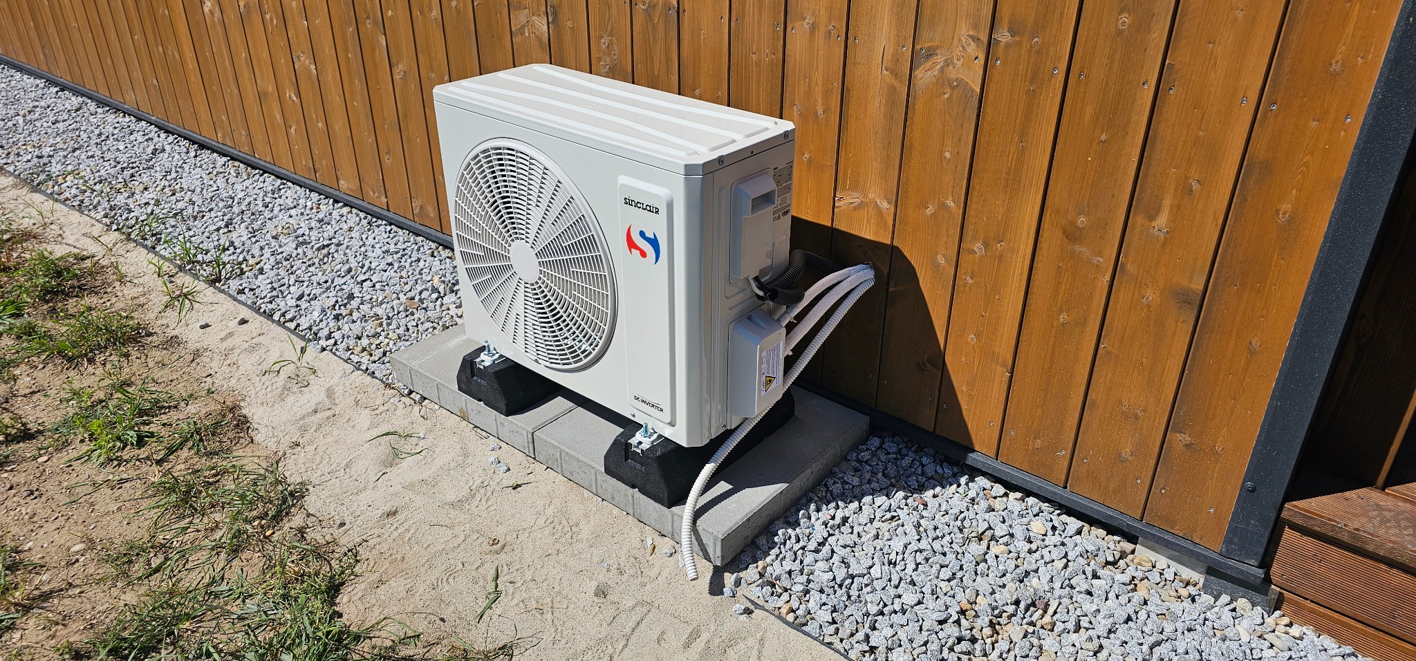 Zewnętrzna jednostka klimatyzacji Sinclair zamontowana na betonowych bloczkach przy drewnianej elewacji budynku, z widocznymi przewodami i drobnym żwirem wokół.