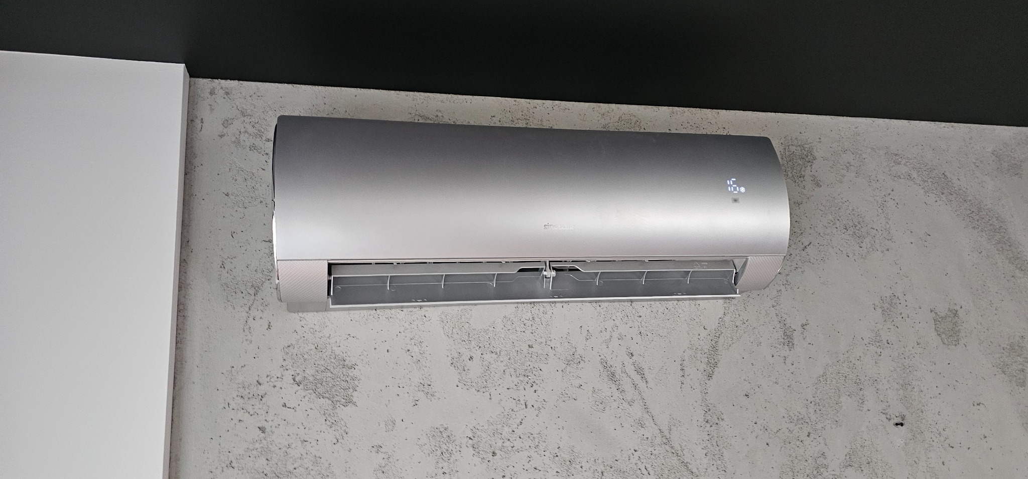 Srebrny klimatyzator Sinclair zamontowany na szarej ścianie o teksturze betonu, widoczny wyświetlacz z temperaturą 16 stopni Celsjusza.