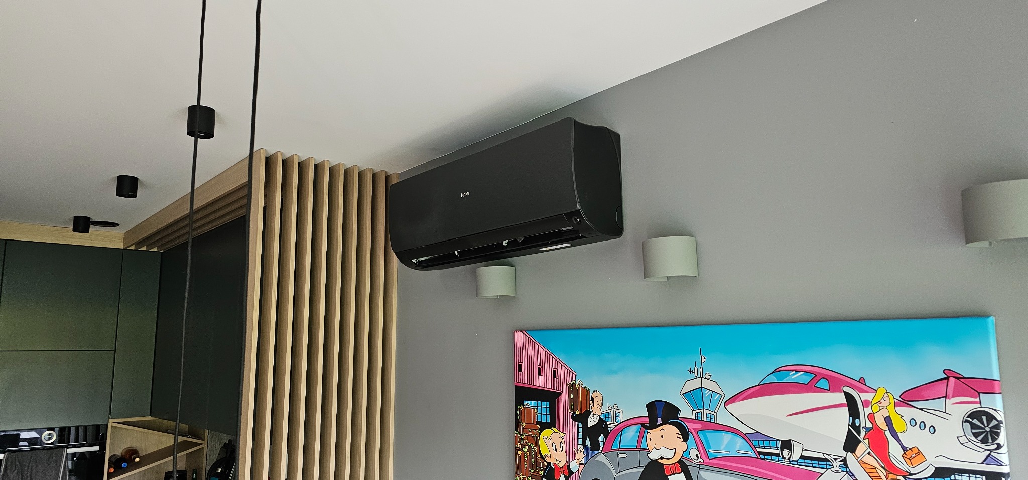 Czarna jednostka klimatyzacji Haier zamontowana na szarej ścianie w nowoczesnym wnętrzu, obok drewnianej ścianki działowej i kolorowego obrazu w stylu pop-art.