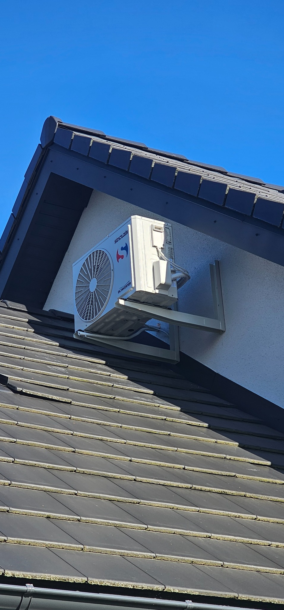 Zewnętrzna jednostka klimatyzacji Sinclair zamontowana na wsporniku na ścianie domu, widoczna na tle ciemnego dachu i błękitnego nieba.