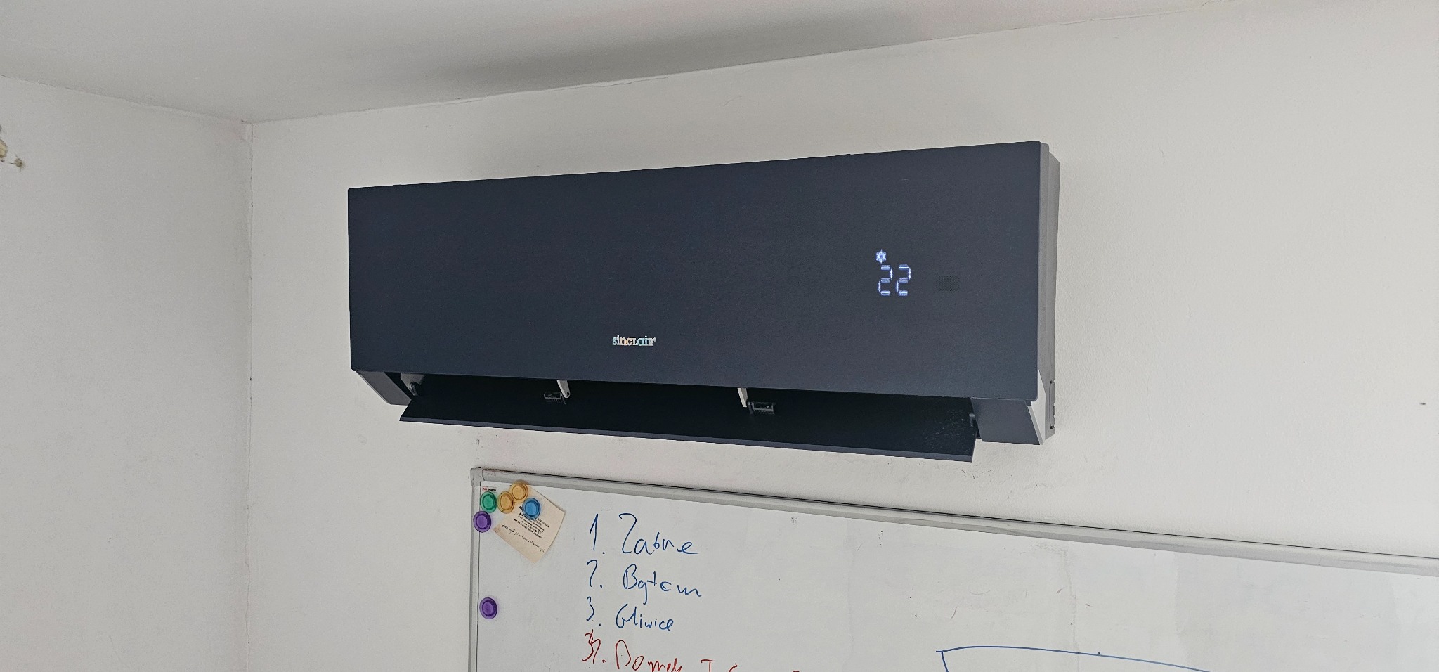 Czarny klimatyzator Sinclair zamontowany na białej ścianie, wyświetlający temperaturę 22 stopnie, pod nim widoczna tablica suchościeralna z zapiskami.