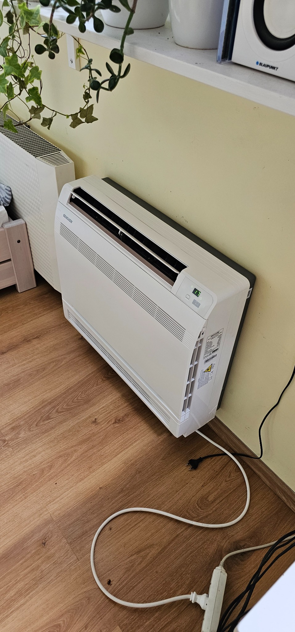 Biały klimatyzator Sinclair zamontowany przy ścianie w pomieszczeniu, widoczne kable zasilające i gniazdko elektryczne na drewnianej podłodze, obok grzejnik i roślina doniczkowa.