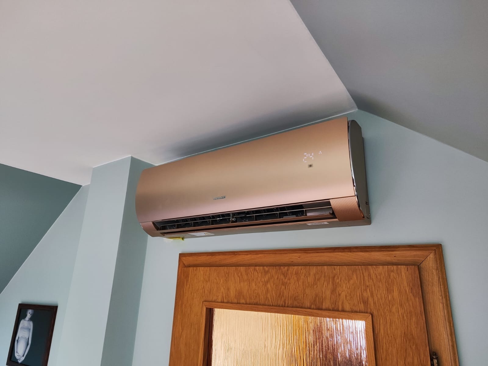 Klimatyzator ścienny w kolorze szampańskiego złota zamontowany nad drewnianymi drzwiami w pokoju z jasnoniebieskimi ścianami i skośnym sufitem.