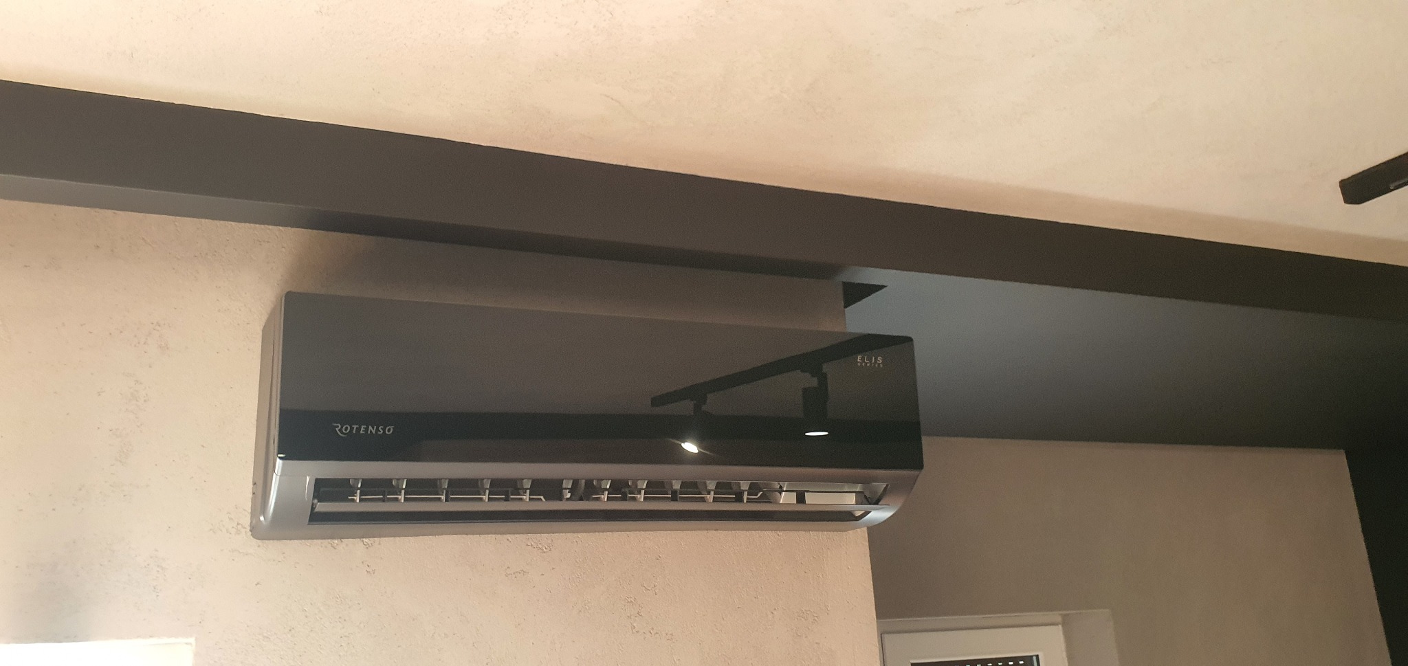 Czarny klimatyzator Rotenso Elis Series zamontowany na ścianie w pomieszczeniu z ciemnymi belkami sufitowymi.