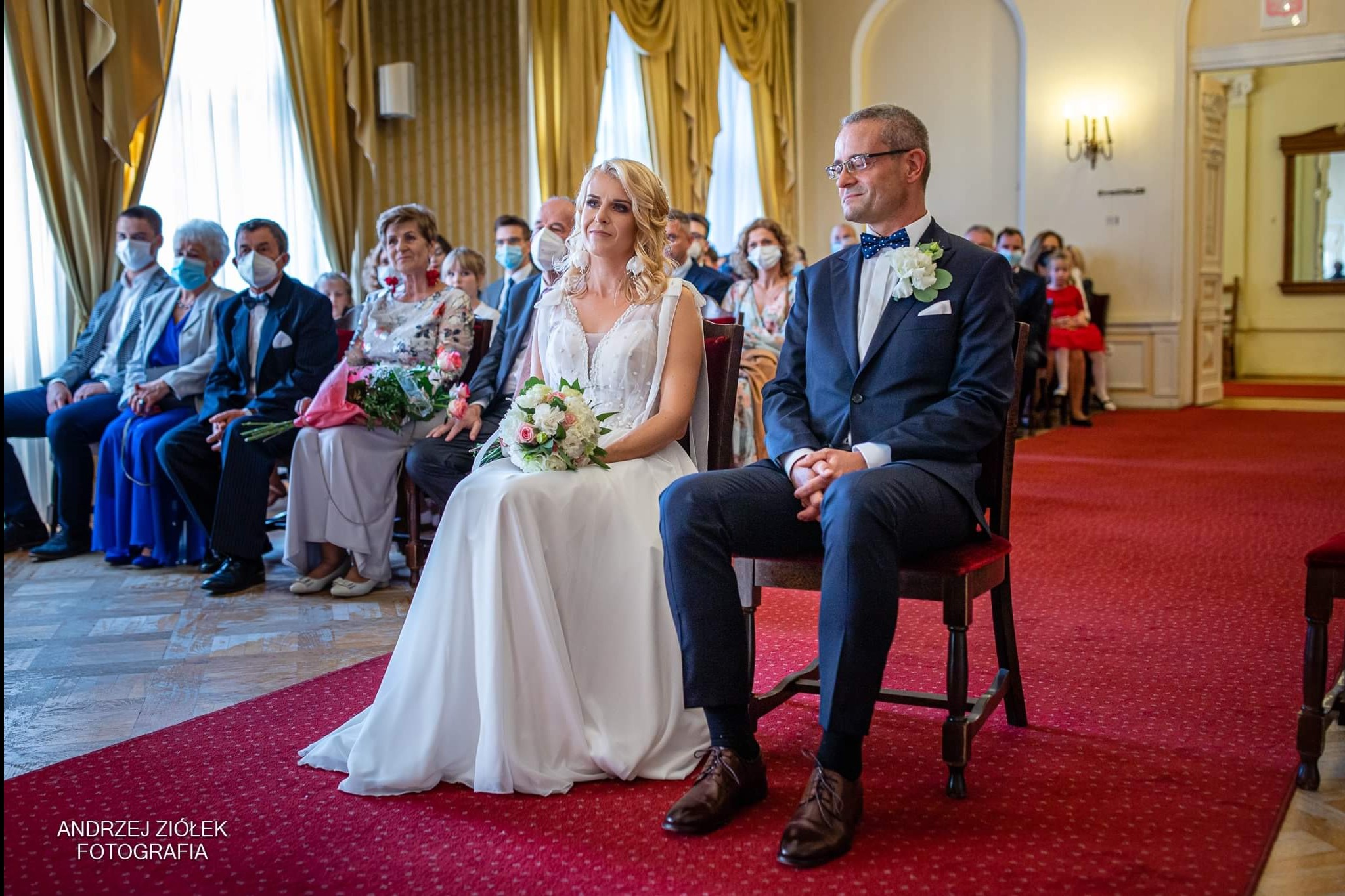 Para młoda siedzi na krzesłach podczas ceremonii ślubnej, goście w tle, czerwony dywan.