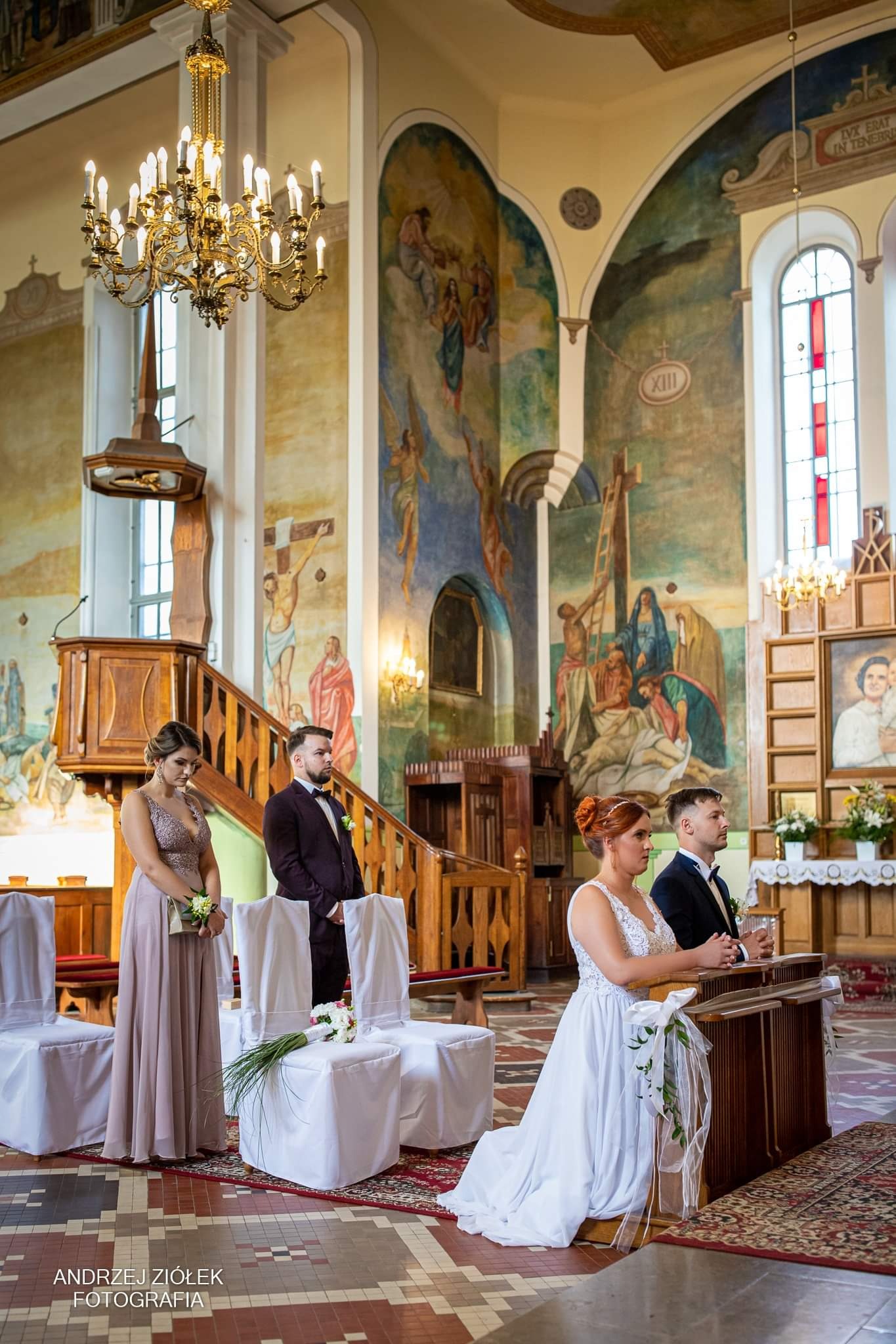 Para młoda klęczy w kościele podczas ceremonii ślubnej, w tle drużba i świadkowa, freski na ścianach i bogato zdobiony żyrandol.