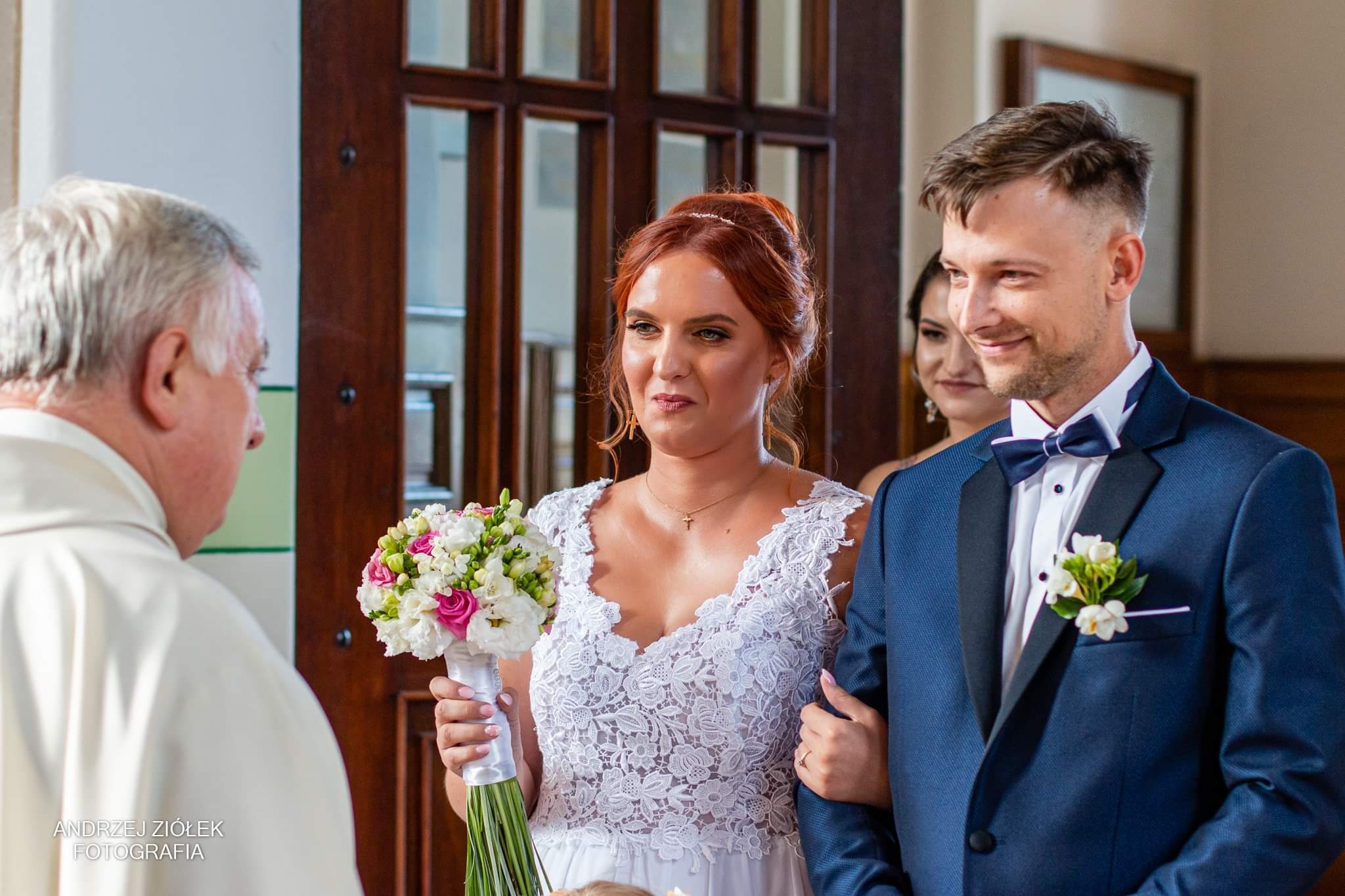 Panna młoda w koronkowej sukni ślubnej trzyma bukiet różowych i białych kwiatów podczas ceremonii zaślubin, obok stoi pan młody w granatowym smokingu z muszką.