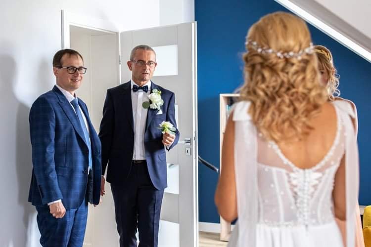 Panna młoda w białej sukni ślubnej, widziana od tyłu, z dwoma elegancko ubranymi mężczyznami w garniturach czekającymi w tle, jeden z butonierką.