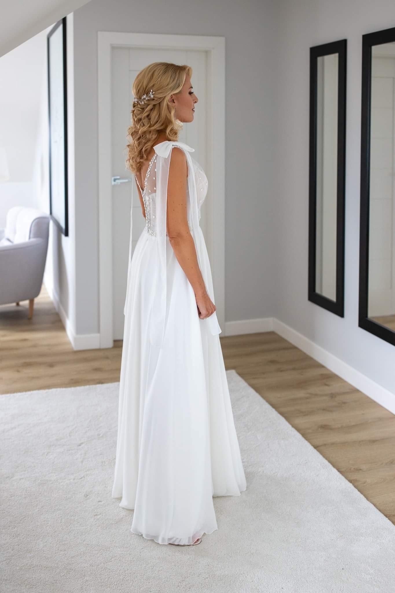 Kobieta w białej sukni ślubnej z delikatnymi, przezroczystymi rękawami i upiętymi blond włosami z ozdobną spinką, pozująca bokiem w jasnym wnętrzu z dwoma lustrami w czarnych ramach.