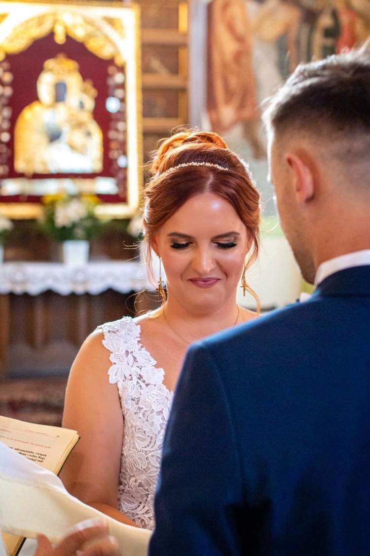 Panna młoda w białej koronkowej sukni, z upiętymi rudymi włosami i delikatnym makijażem, spogląda z uśmiechem na pana młodego podczas ceremonii w kościele. W tle widać złoty obraz i kwiaty.