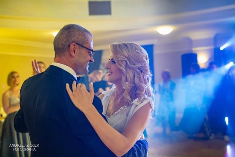 Para młoda podczas pierwszego tańca, mężczyzna w czarnym smokingu i okularach, kobieta w białej sukni z upiętymi blond włosami, uśmiechnięci patrzą na siebie, w tle goście i oświetlenie sceniczne.