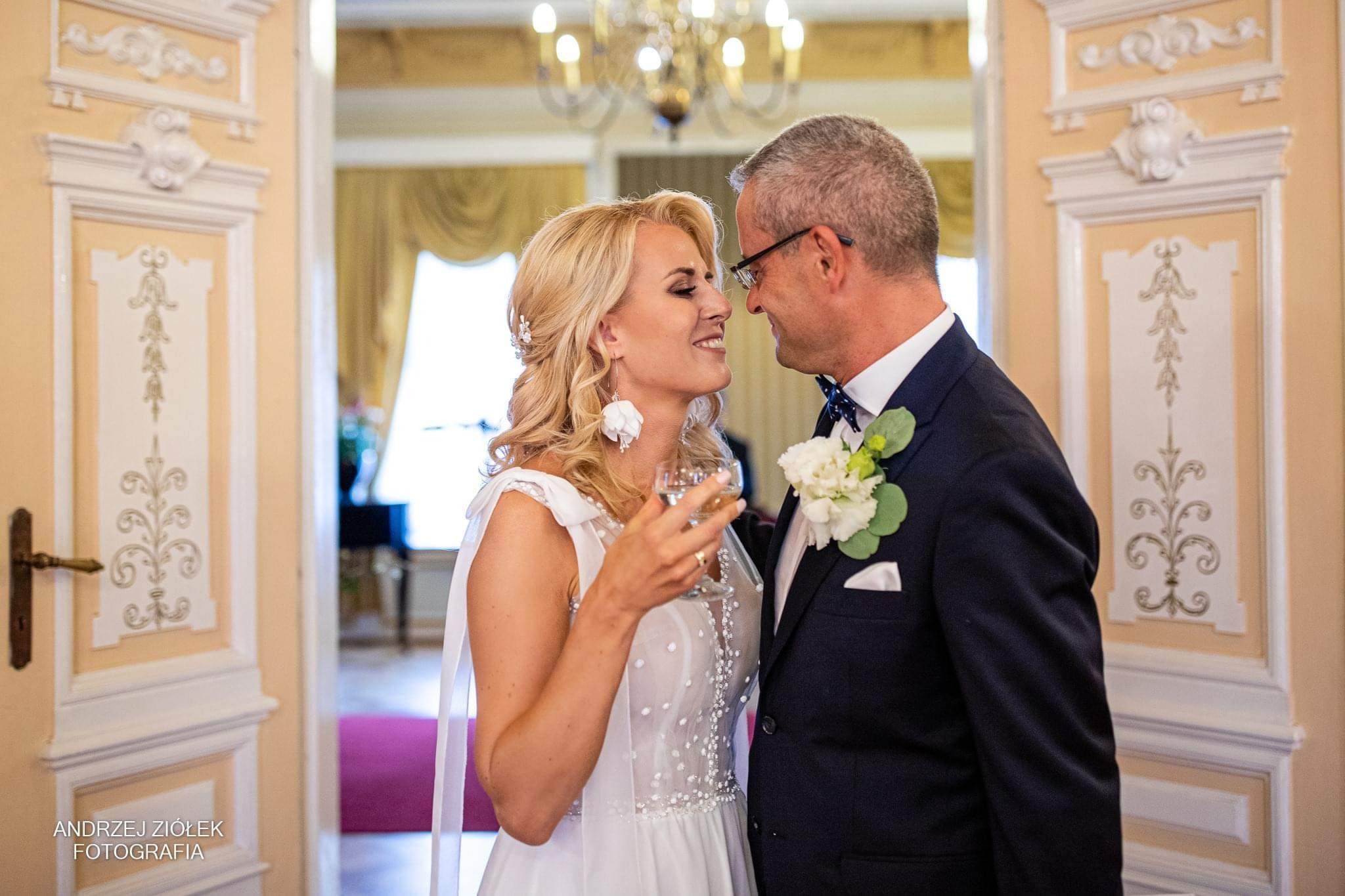 Uśmiechnięta panna młoda z blond włosami i kwiatowymi kolczykami, trzymająca kieliszek wina, patrzy na pana młodego w granatowym garniturze z butonierką, na tle klasycznego wnętrza z żyrandolem