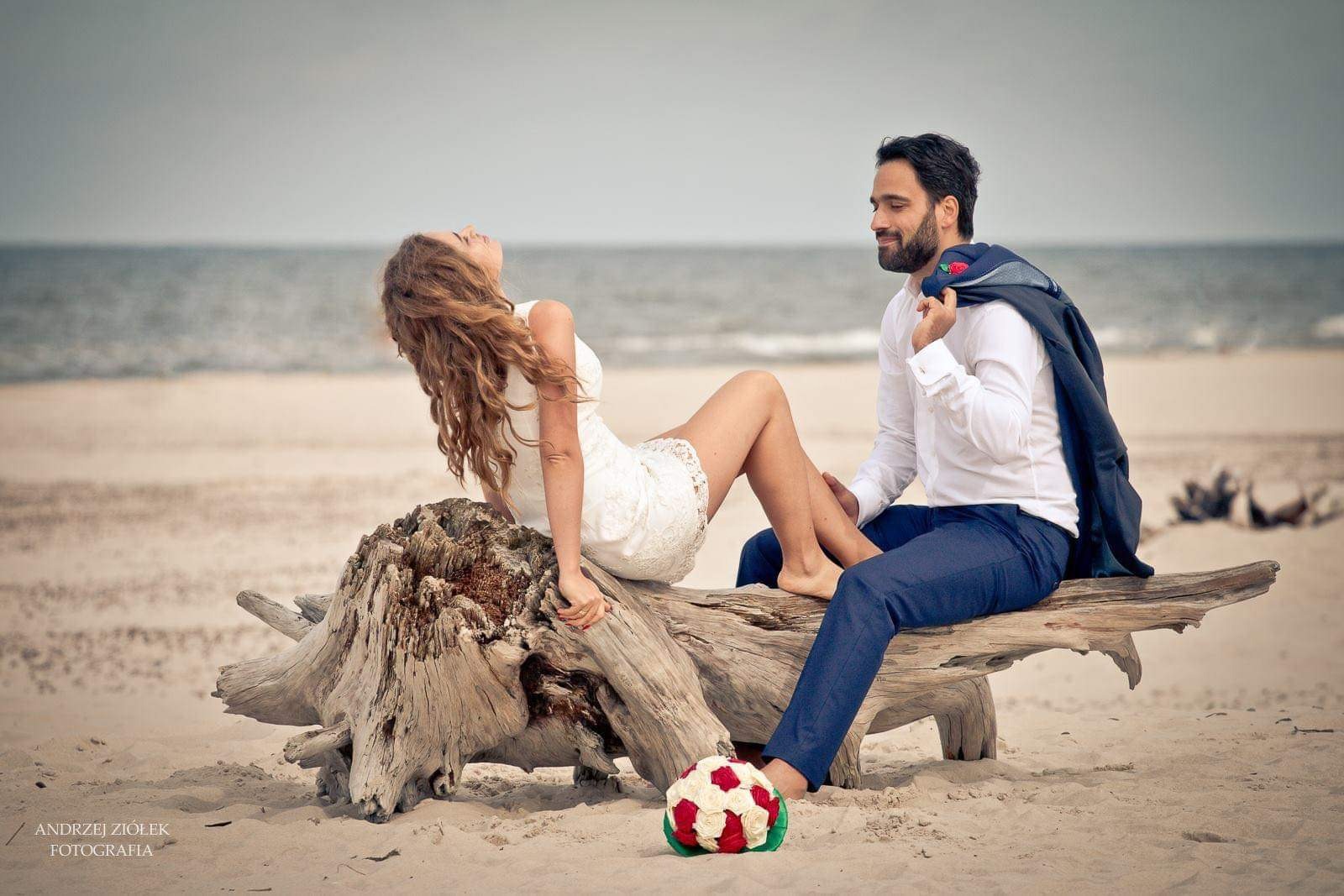 Para młoda siedząca na pniu drzewa na plaży, ona w koronkowej sukience, on w białej koszuli i granatowych spodniach, z marynarką na ramieniu, bukiet róż leży na piasku, w tle morze.