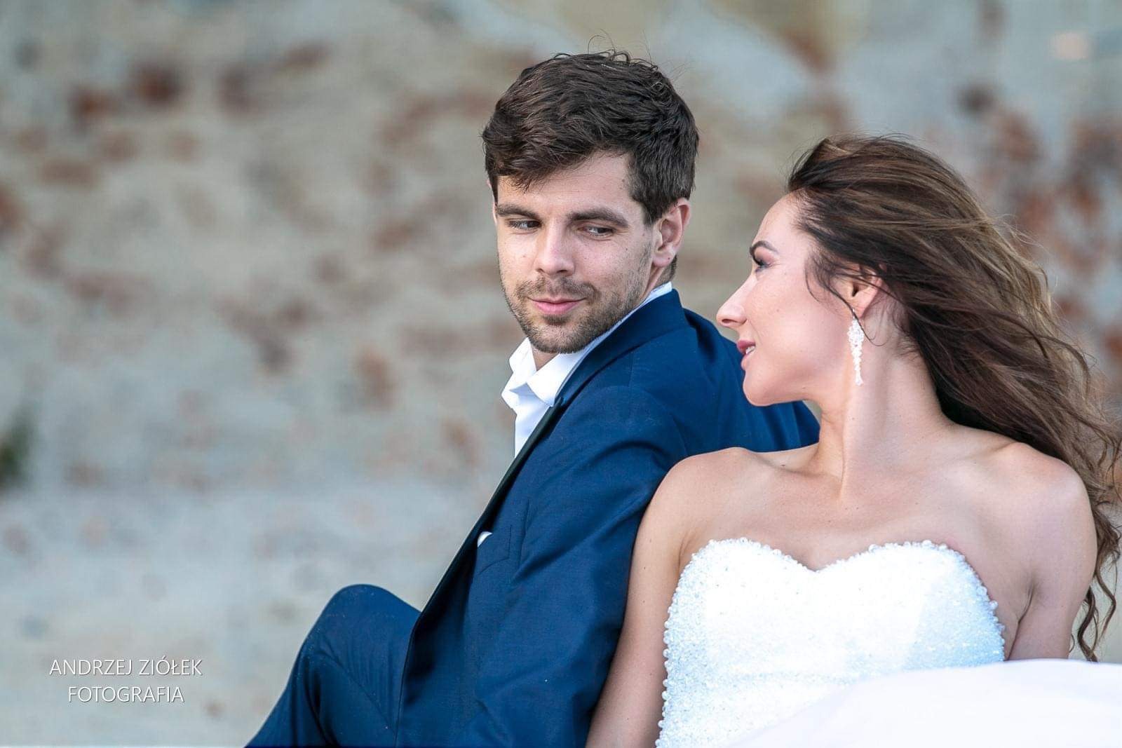 Elegancka para młoda w plenerze: pan młody w granatowym garniturze spogląda czule na uśmiechniętą pannę młodą w białej sukni bez ramiączek z błyszczącymi aplikacjami, w tle ceglana ściana.
