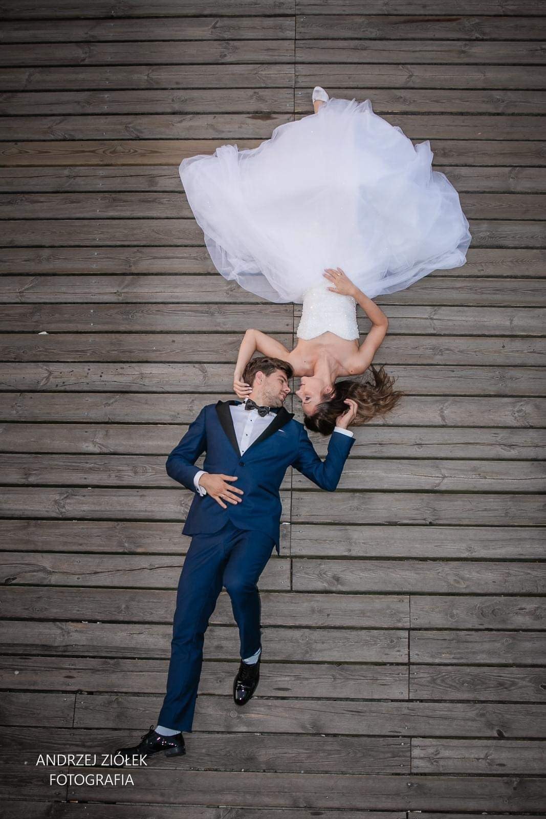 Para młoda leżąca na drewnianym pomoście, panna młoda w białej sukni, pan młody w granatowym garniturze z muszką, spojrzenia skierowane ku sobie.
