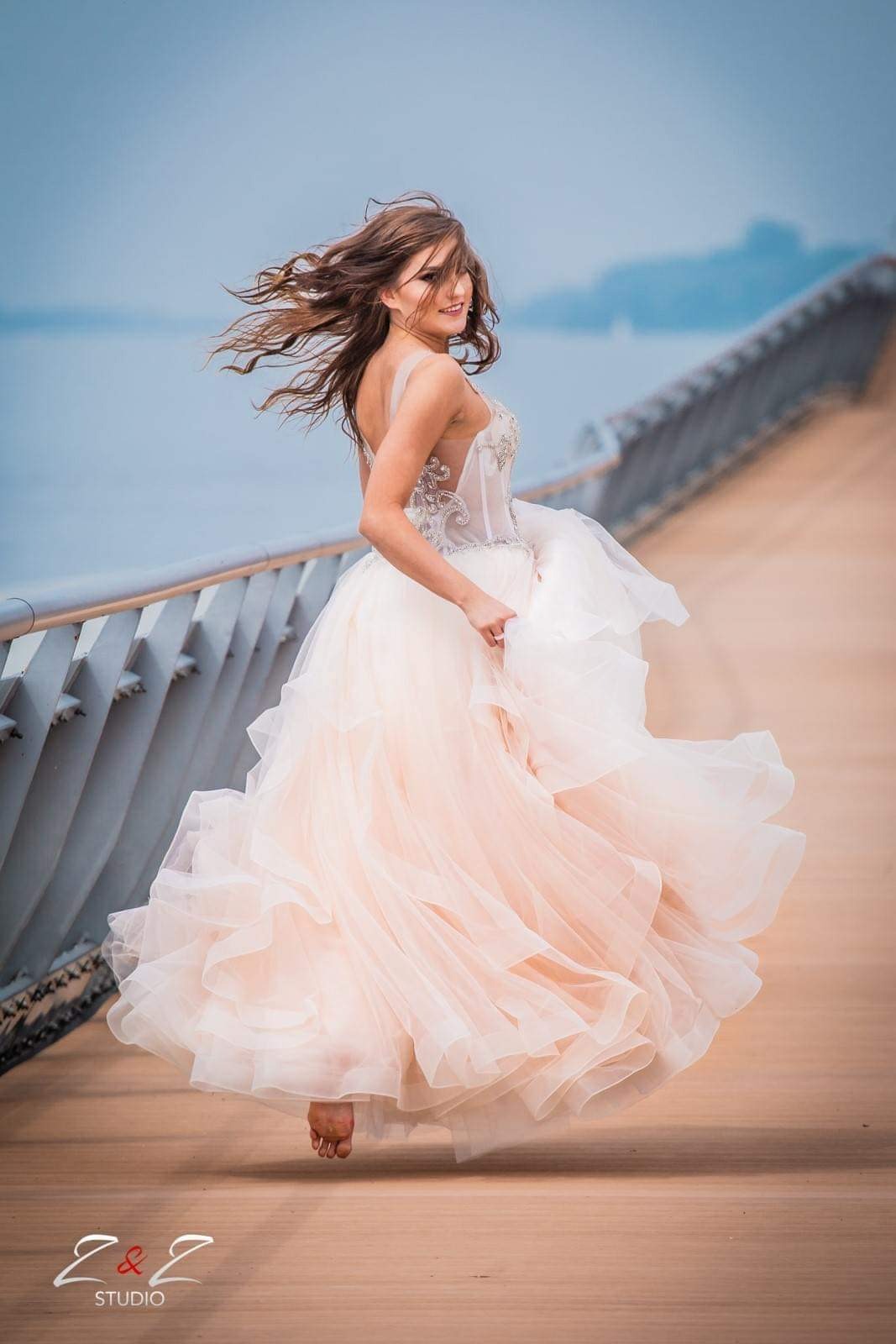 Młoda kobieta w zwiewnej, warstwowej sukni balowej w odcieniach pudrowego różu i bieli, biegnąca boso po drewnianym pomoście z metalową barierką, z wiatrem we włosach i uśmiechem spoglądająca przez...