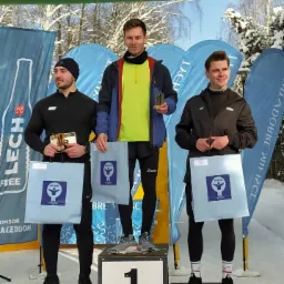Trzech mężczyzn na podium po biegu zimowym, trzymających nagrody i torby sponsorskie. Pierwszy zawodnik stoi na najwyższym stopniu, w tle śnieg i baner startowy.