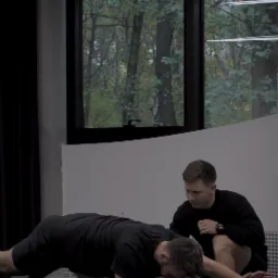 Mężczyzna wykonuje ćwiczenie na macie pod nadzorem drugiego mężczyzny, w tle okno z widokiem na drzewa jesienią.