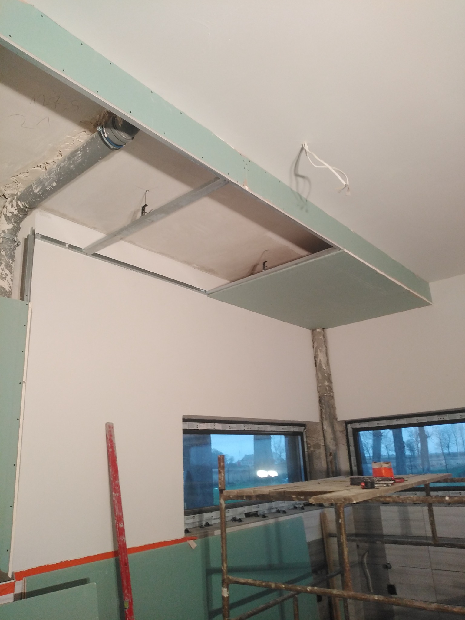 Wnętrze w trakcie montażu podwieszanego sufitu z płyt gipsowo-kartonowych, widoczne metalowe profile, rury instalacyjne i fragment ściany z oknami, z zewnątrz widok na drzewa.
