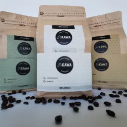 Trzy opakowania kawy ziarnistej marki PiKawa na białym tle, z rozsypanymi ziarnami kawy. Opakowania w odcieniach brązu i zieleni, z widocznym logo i informacją o blendzie 100% Arabica...