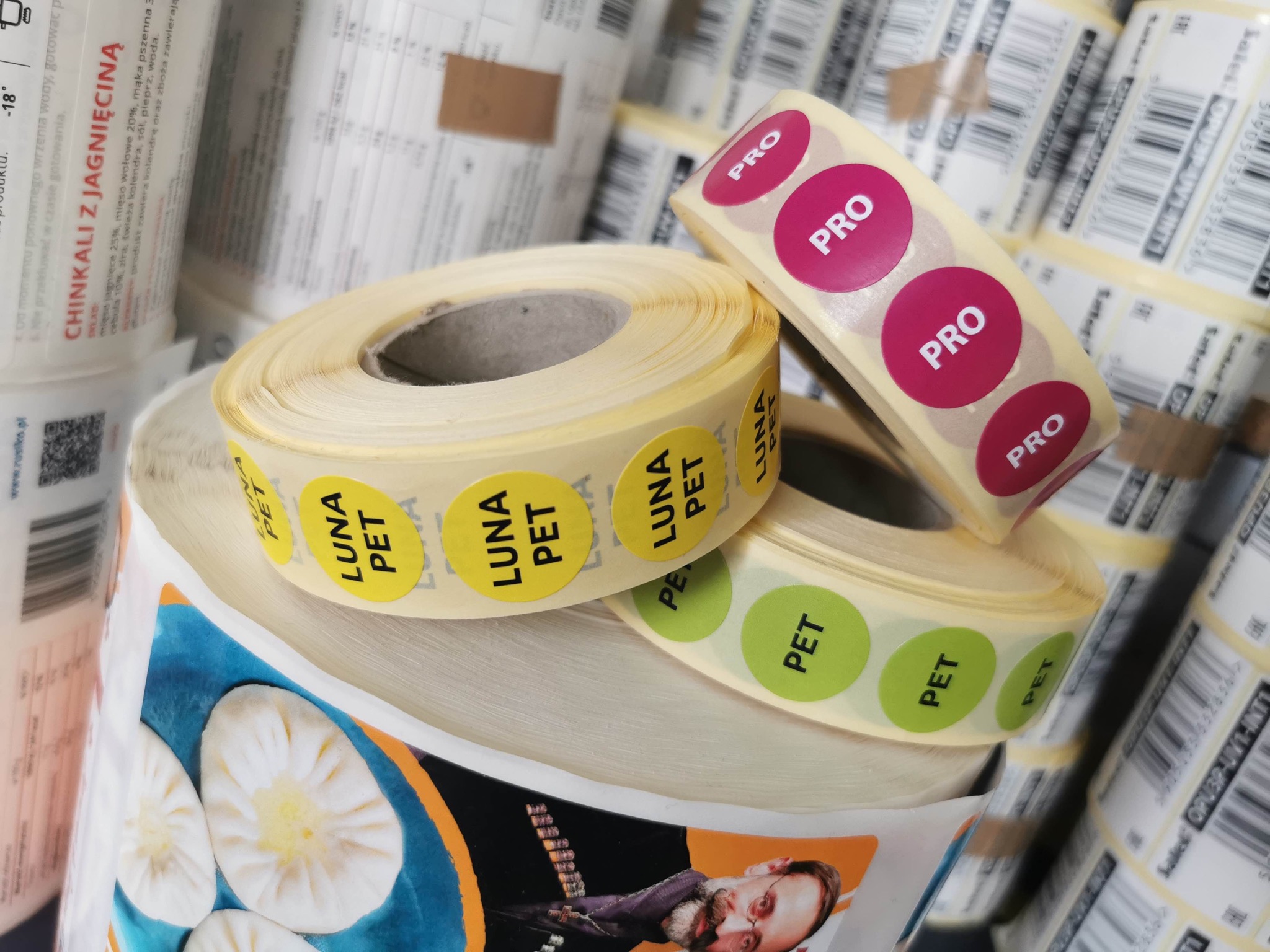 Różne rolki okrągłych etykiet samoprzylepnych z nadrukami 'LUNA PET' w kolorach żółtym i zielonym oraz 'PRO' w kolorze różowym, ułożone na stosie opakowań z nadrukiem.