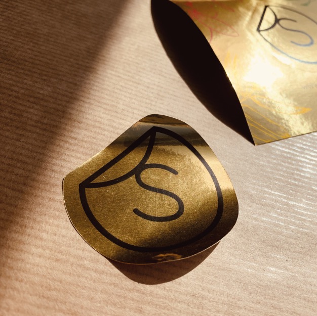 Złota naklejka w kształcie łezki z czarnym logo litery S, częściowo odklejona od podłoża, obok arkusz złotej folii z odręcznym rysunkiem, oświetlona ciepłym światłem.