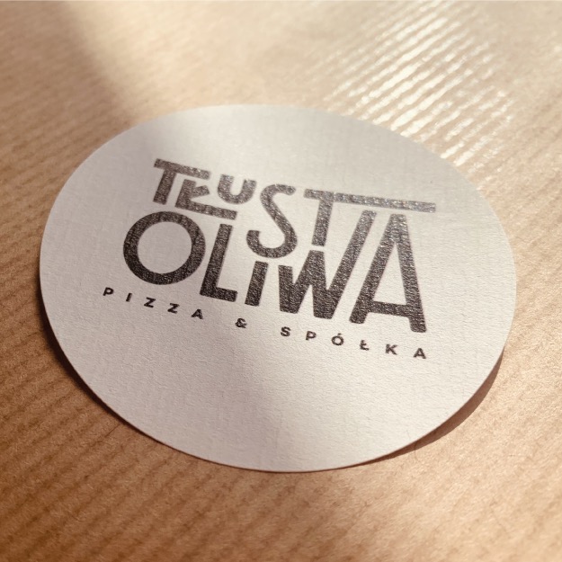 Okrągła naklejka z logo 'Tłusta Oliwa Pizza & Spółka' na brązowym papierze, oświetlona naturalnym światłem z góry, tworząca cień na powierzchni naklejki.