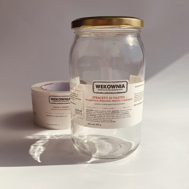 Pusta szklana słoik z etykietą 'Stracetti di Filetto - Poledwica Wołowa Prosto z Bawełny' obok rolki etykiet 'Syrop z Pigwy' marki Wekownia, w jasnym oświetleniu.