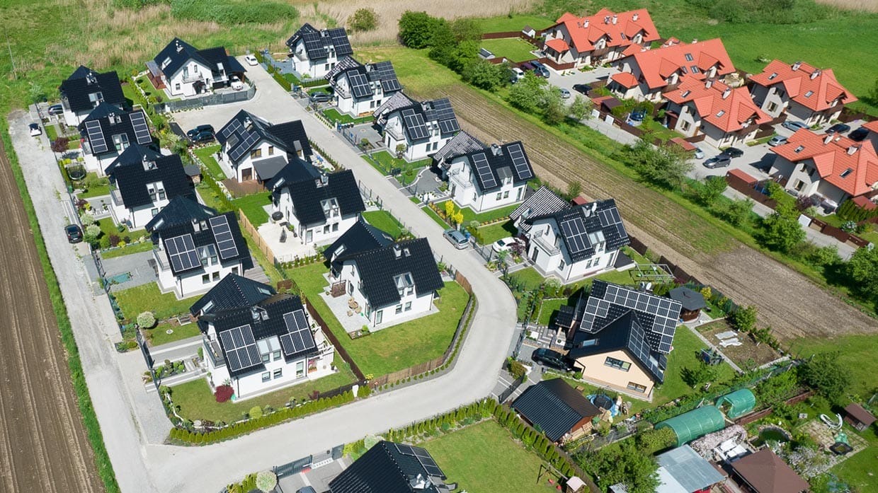 Widok z lotu ptaka na osiedle domów jednorodzinnych z panelami słonecznymi na dachach, część domów z czerwonymi dachami, a pozostałe z czarnymi, otoczone zielenią i polami.