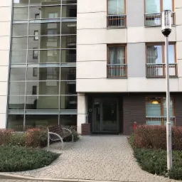 Nowoczesny budynek biurowy z przeszkloną fasadą, brukowanym chodnikiem i ławką na zewnątrz.