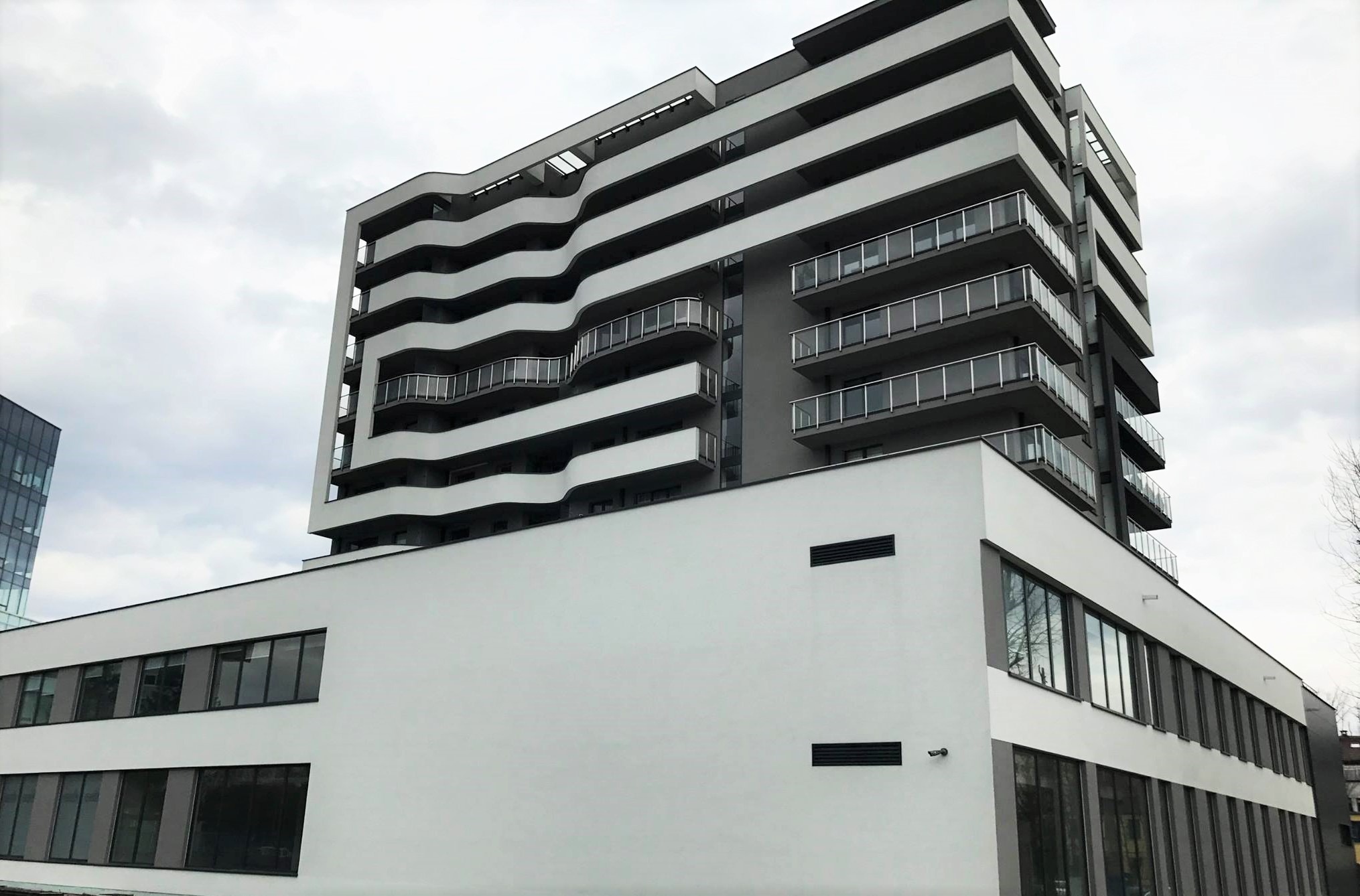 Nowoczesny budynek mieszkalny z balkonami o falistym kształcie, widok z dołu na tle pochmurnego nieba.