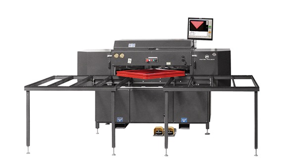 Duże, szare urządzenie przemysłowe z czerwonym elementem w centrum i monitorem na górze, z dwoma metalowymi stołami po bokach, na białym tle.