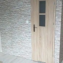 Wnętrze pokoju z dekoracyjną ścianą imitującą kamień, drewnopodobną podłogą i drzwiami z przeszkleniami, widoczne z niskiego kąta.