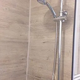 Nowoczesny zestaw prysznicowy z deszczownicą i słuchawką, zamontowany na ścianie wyłożonej płytkami imitującymi drewno w jasnym odcieniu, widoczne elementy armatury i kabina prysznicowa.