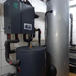 Zamknięte pomieszczenie techniczne z zainstalowanym systemem grzewczym: szara jednostka sterująca z podświetlonym logo i ekranem, dwa zbiorniki akumulacyjne (szary i srebrny) oraz rury z izolacją...